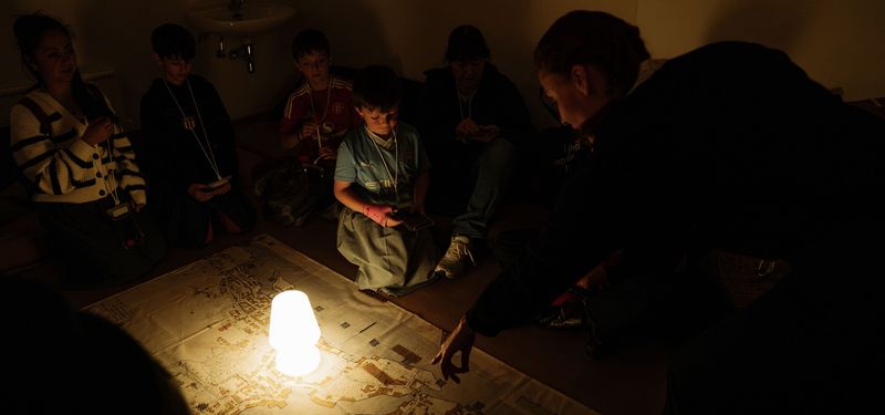 A dimly lit room with children sat on the floor around a fabric map. The actor points to a location on the map and the audience uses phones to scan this location