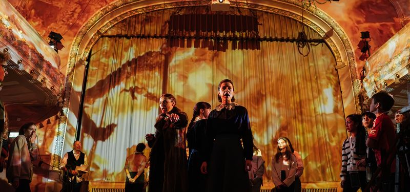 Inside a theatre space, the actors are in the centre surrounded by the audience while the walls and roof of the building appear to be consumed by flames