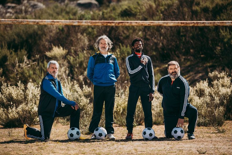 Photograph of four actors in football outfits, in front of a rusting goal