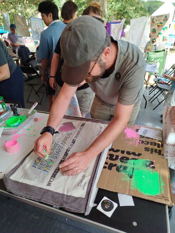 A person wearing a hat and glasses leans over their tea towel, mopping up a small spill of bright green ink