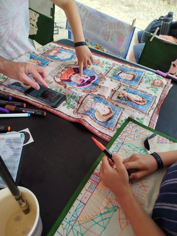 A table covered in craft materials, two people using fabric pens to draw on the back of tea towels