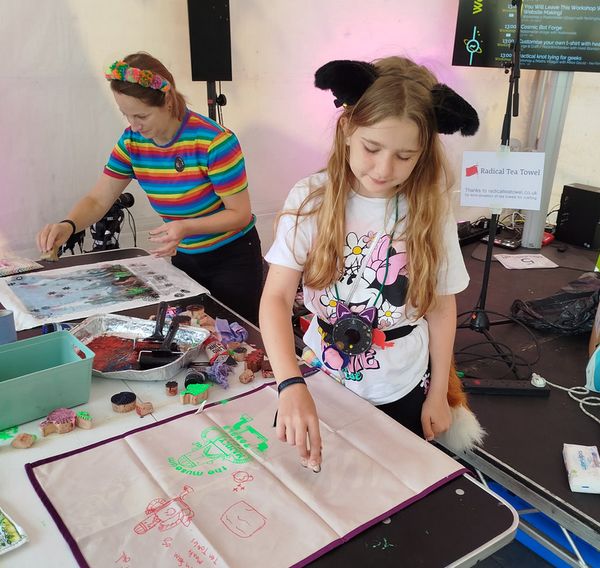 A person wearing a rainbow t-shirt and a young person with cat ears, using stamps to decorate tea towels.