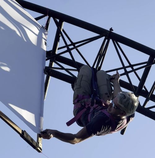 Photograph of a theatre technician hanging upside down from a giant truss and fixing a projection screen.