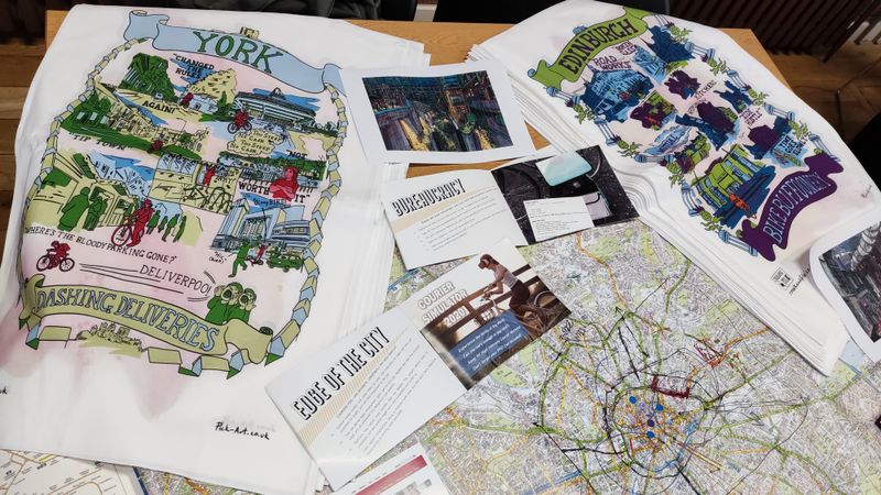 A photograph of a table with tea towels, booklets and maps on it, with a bicycle in the background. The tea towels are designed to reflect the experiences of cycle couriers in different cities.